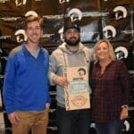 Three people standing in front of a Supreme Boats banner with a plaque.