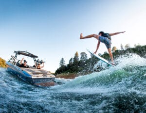 Person falls off a surfboard while wake surfing behind a Supreme Towboats S220, with others on the boat watching and taking photos. Water splashes and trees are visible in the background.