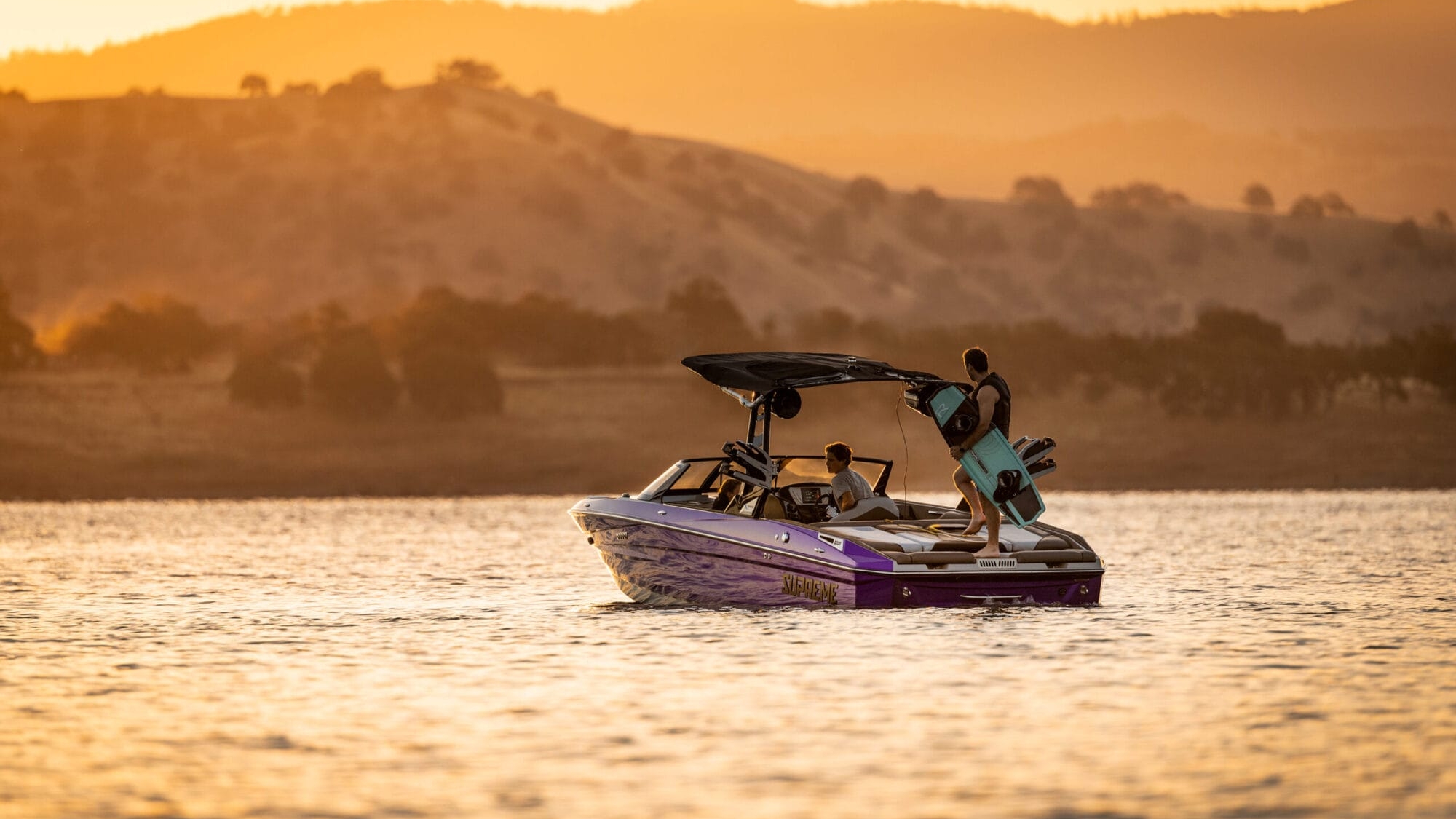 A person holding a wakeboard stands on a 2026 Supreme S220 purple boat in a lake at sunset, with hills and trees in the background.