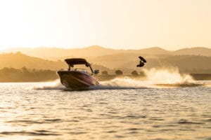 A person wakeboarding is airborne behind a 2026 Supreme Towboats S220 on a lake during sunset, with mountains visible in the background.