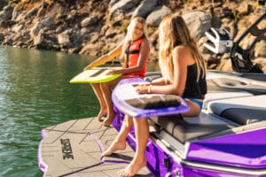 Two girls with surfboards sit on the back of a purple Supreme S220 near a rocky shore; one relaxes on the deck while the other smiles from a seat, both enjoying their Supreme Towboats S220 adventure.