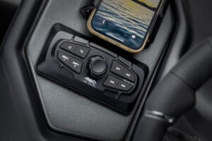 Close-up of a Supreme S220 boat dashboard audio control panel with various buttons, a central dial, and a smartphone resting above it.