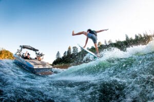A person in colorful shorts surfs on a wave behind a Supreme Boats S240, with two people watching from the boat and trees visible on the shore in the background.
