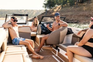 Four people in swimsuits sit and relax on a 2026 Supreme S240 wakeboat, smiling and talking while on a lake with dry, hilly landscape in the background.