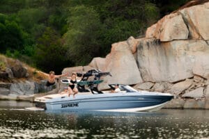 Three people on a 2026 Supreme Boats S240 by rocky cliffs, with two jumping into the water and one seated at the edge, while another person drives the boat.