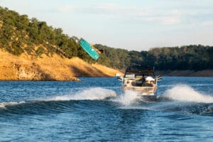 A person wakeboarding behind a 2026 Supreme Boats S240 on a lake, with grassy hills and trees in the background beneath a clear sky.