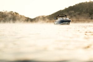 A 2026 Supreme S240 motorboat floats on calm water with mist rising, surrounded by tree-covered hills in the background.