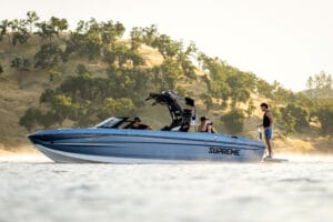 A blue 2026 Supreme Boats S240 speedboat with three people on board is stationary on a calm lake, surrounded by trees and hills in the background.