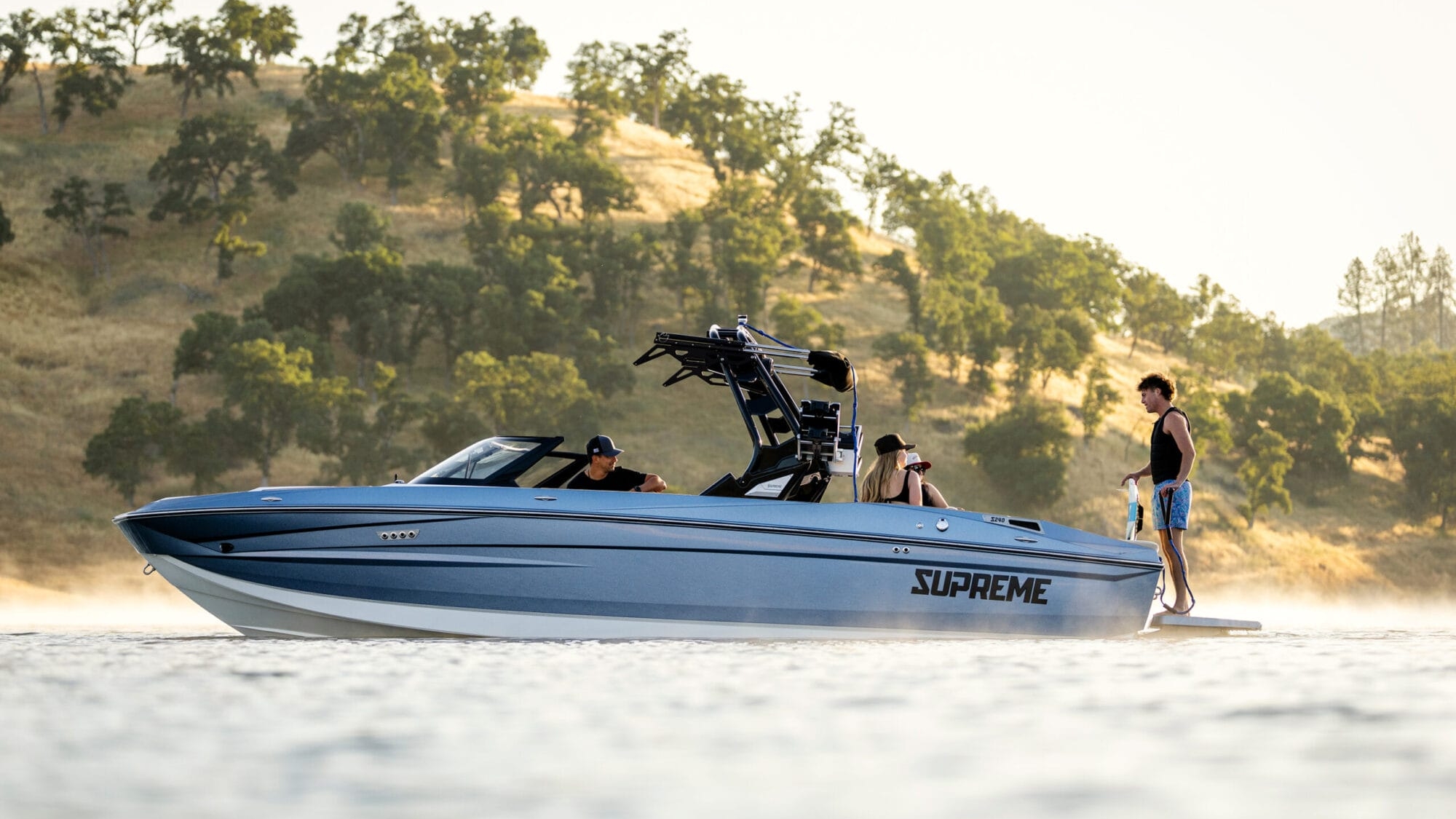 A blue 2026 Supreme Boats S240 speedboat with three people on board is stationary on a calm lake, surrounded by trees and hills in the background.
