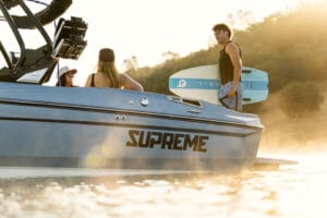 Three people are on a 2026 Supreme Boats S240 in the water; one person holds a surfboard as the sun sets behind the trees.