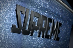 The word "SUPREME" in bold, black, three-dimensional letters is displayed against a blue, glittery background, evoking the sleek style of the Supreme S240.