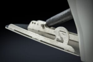 Close-up view of a metal ice skate blade attached to a white boot, showcasing detailed construction and a cut-out letter "S" in the blade holder, reminiscent of the sleek design seen on the Supreme S240.