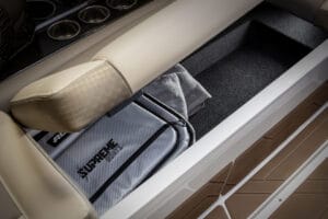 A storage compartment under a beige Supreme S240 boat seat contains a grey bag labeled "Supreme Boats" and a grey towel. Cup holders and flooring are also visible.