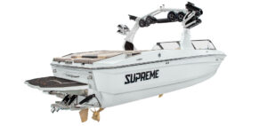 White Supreme S220 wakeboard boat with “SUPREME” lettering, tan seats, and a tower equipped with speakers and board racks, viewed from the rear side.