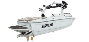 A white Supreme Towboats S220 wakeboard boat with tan seating, a black swim platform, and a tower equipped with speakers and racks, viewed from the rear side angle.