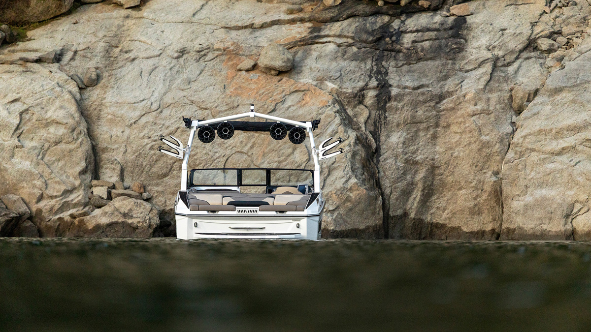 A white Supreme S220 wakeboard boat floats near a rocky cliff, facing the camera with empty seats and speakers mounted on the frame.