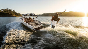 A person wakesurfs behind a white Supreme S220 motorboat on a sunlit lake, leaping above the water with one hand raised while others ride in the boat.