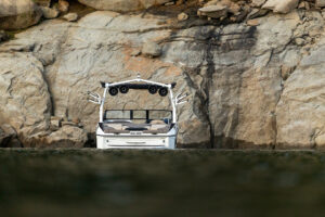 A white Supreme S220 boat with speakers is floating on calm water near a rocky shoreline with large boulders in the background.