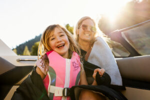 A smiling child in a pink life jacket sits next to an adult woman wearing sunglasses and a blanket on a Supreme S220 boat in sunny weather.