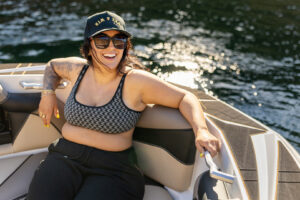 Woman wearing sunglasses and a cap sits smiling on a Supreme S220 boat, with sunlight reflecting on the water behind her.