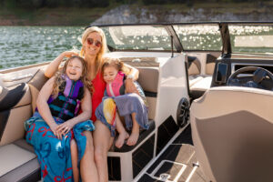 A woman and two children sit on a Supreme S220 boat. The children are wrapped in towels and wearing life jackets, smiling. The boat is on a body of water, with sunlight reflecting on the surface.