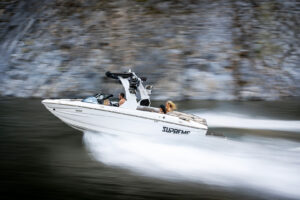A white Supreme S220 speedboat moves quickly across the water near a rocky shoreline, with two people onboard.