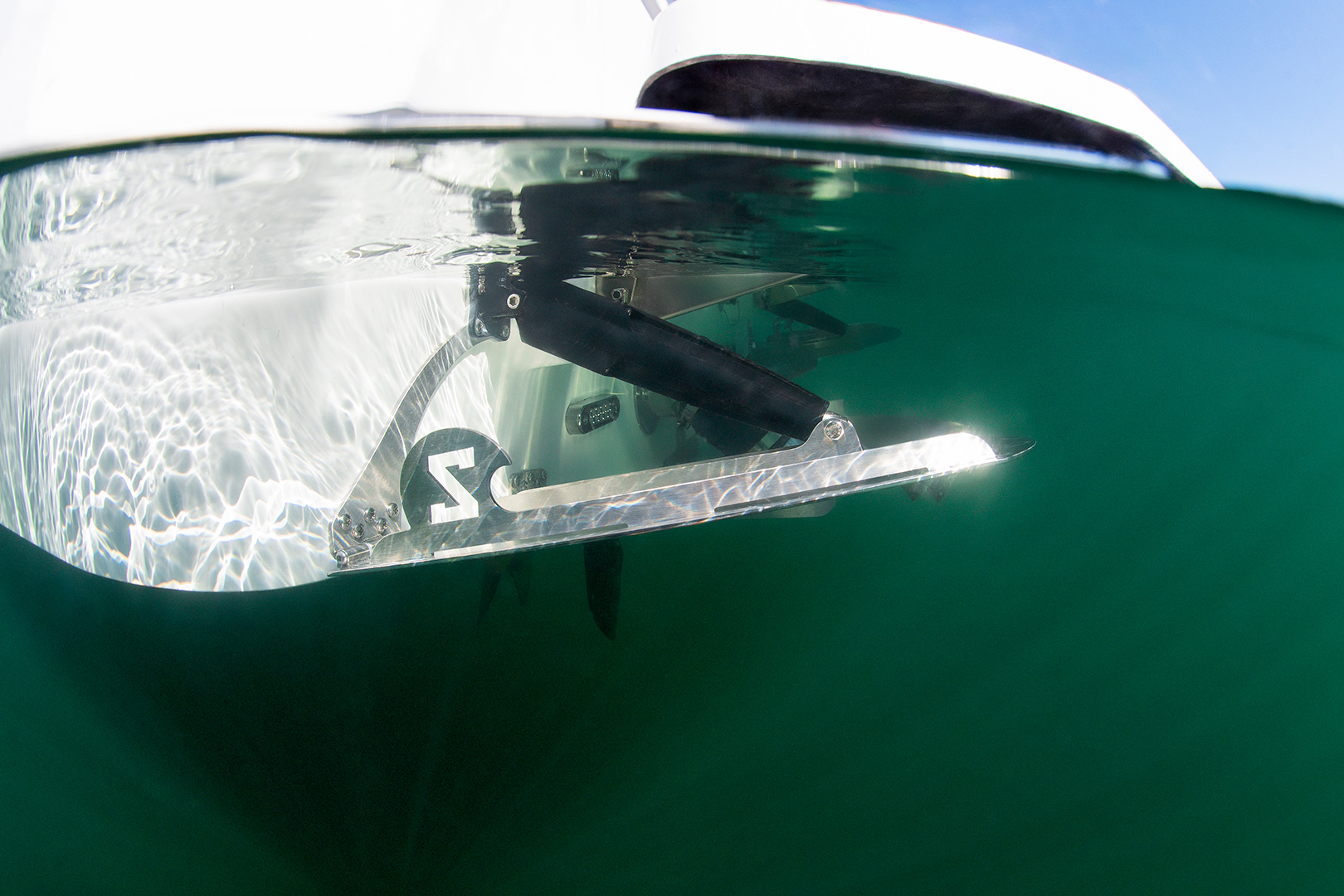 A close-up view of a hydrofoil partially submerged in water, attached to the underside of a Supreme S220, with sunlight reflecting off the metal surface.