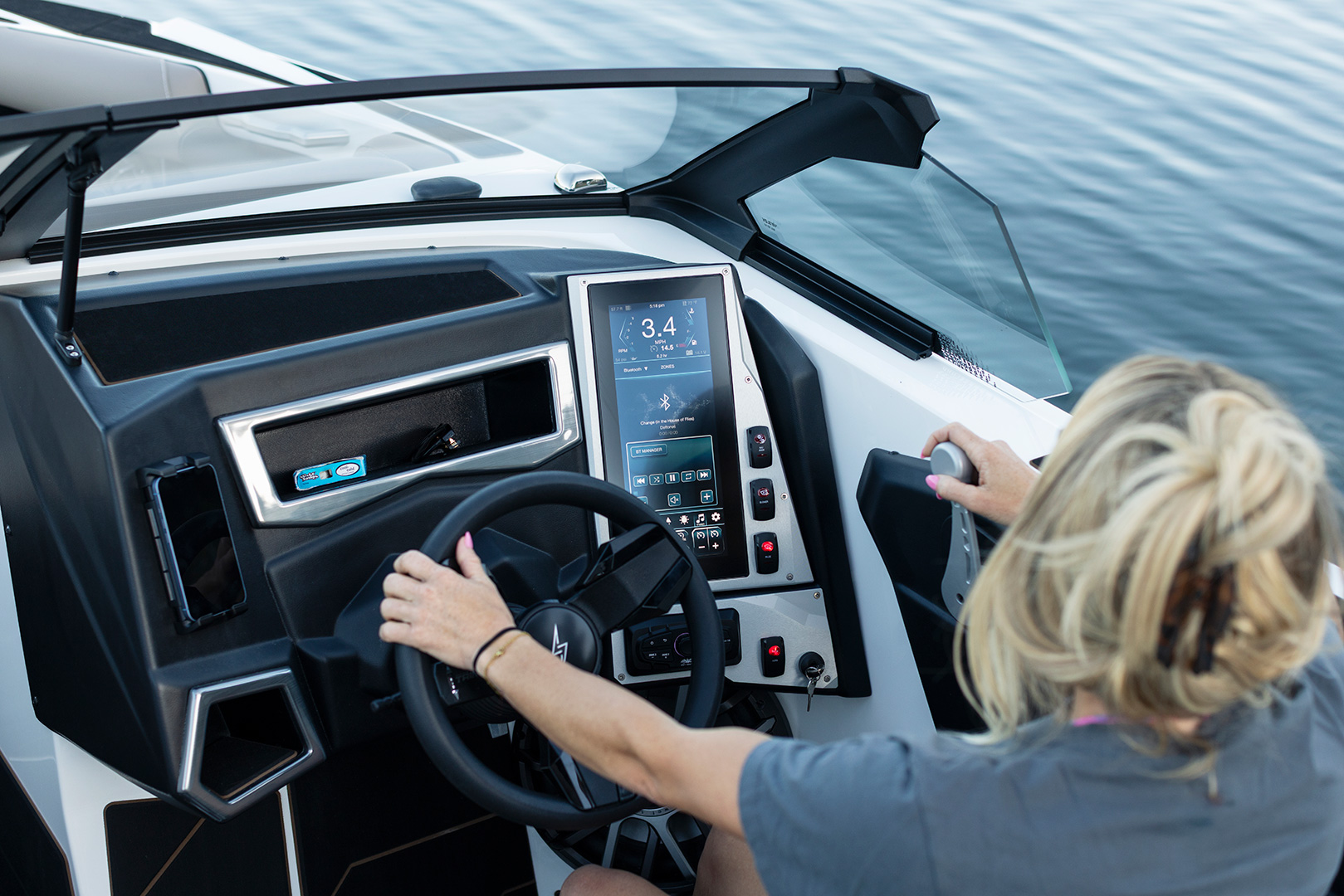 Person steering a modern Supreme Towboats S220, holding the wheel with one hand and a throttle lever with the other, with a digital control panel displayed, on calm water.