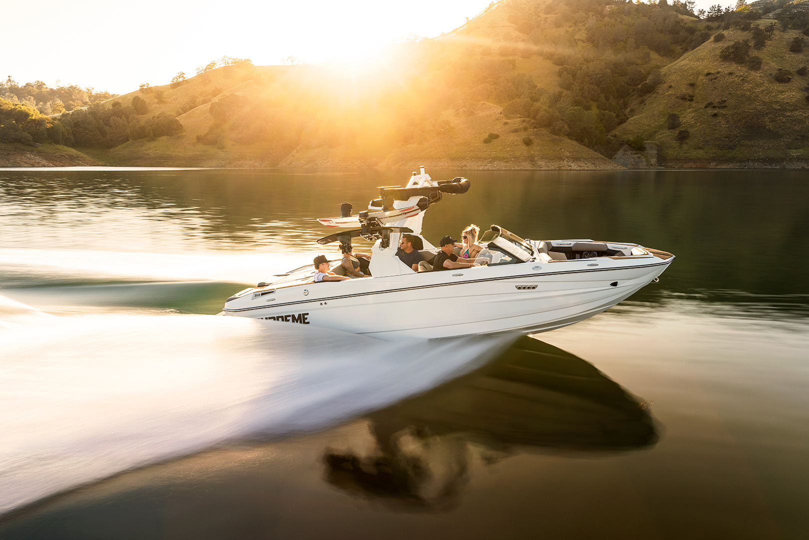 A white Supreme Towboats S220 speedboat with several people onboard moves quickly across a calm lake at sunset, creating a trail in the water. Hills with green grass are visible in the background.