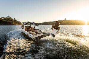 A person wakesurfs behind a white Supreme S220 motorboat on a lake during sunset, creating splashes and waves as the Supreme Towboats S220 speeds forward.
