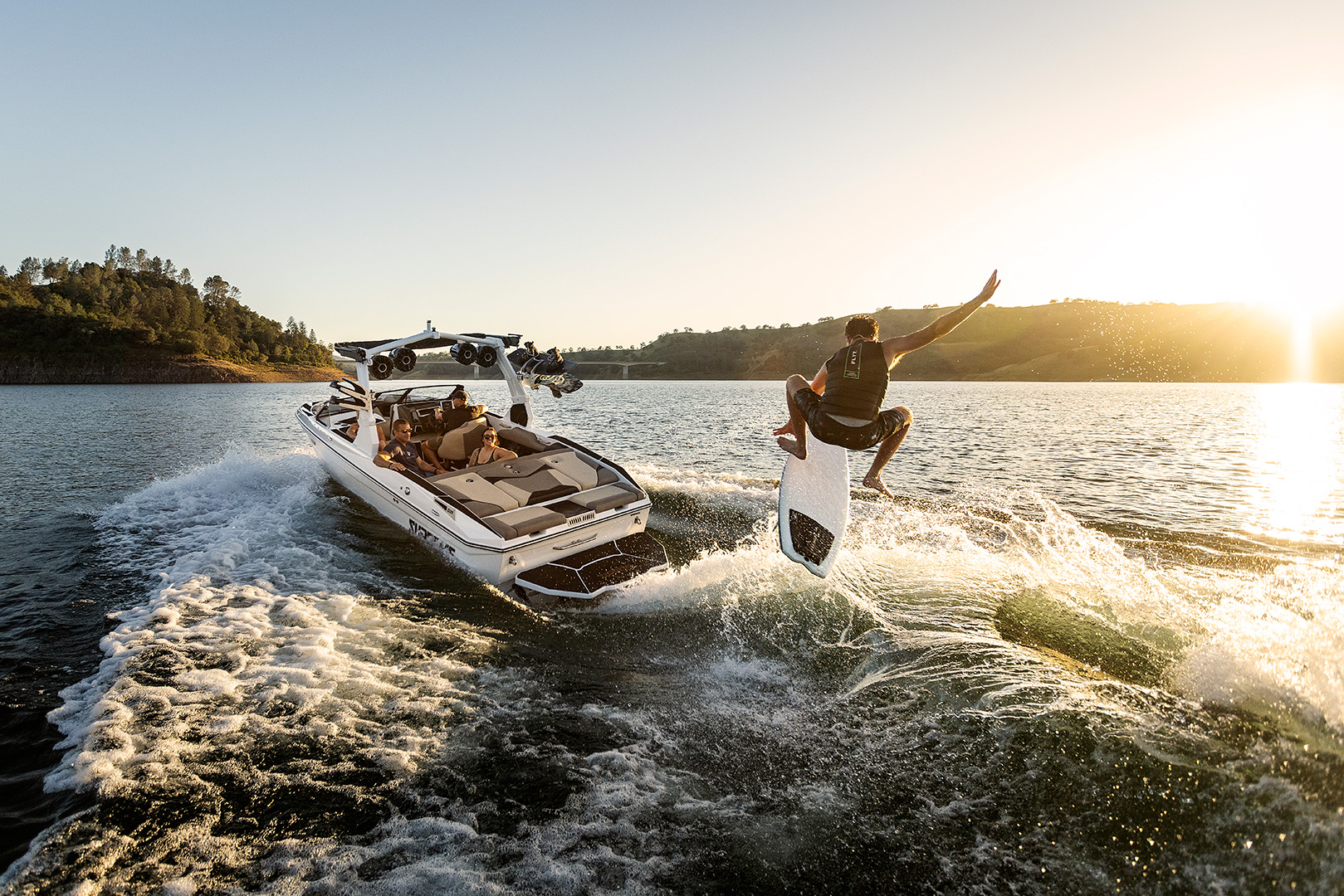 A person wakesurfs behind a white Supreme S220 motorboat on a lake during sunset, creating splashes and waves as the Supreme Towboats S220 speeds forward.