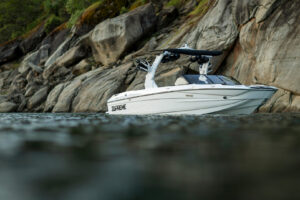 A white Supreme Towboats S220 motorboat with the word “SUPREME” on its side is floating near rocky cliffs and calm water, with greenery visible at the top of the rocks.