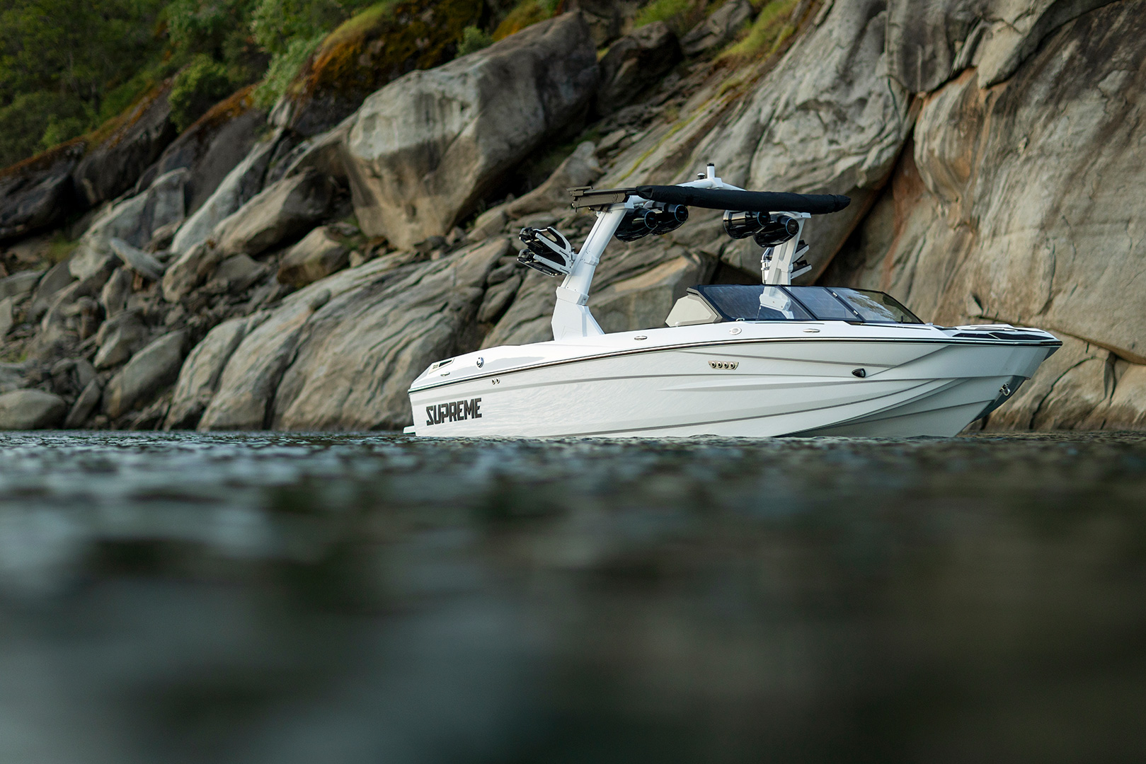 A white Supreme Towboats S220 motorboat with the word “SUPREME” on its side is floating near rocky cliffs and calm water, with greenery visible at the top of the rocks.