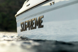 Close-up of a boat partially in water with the word “SUPREME” in bold letters on its side, capturing the sleek design of the Supreme S220. Trees and land are blurred in the background, highlighting the dynamic presence of this Supreme Towboats S220 model.