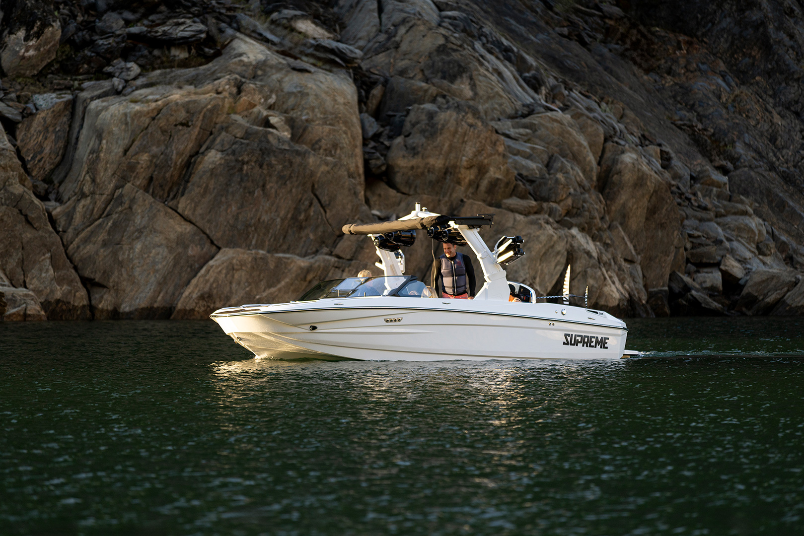 A white Supreme Towboats S220 speedboat with passengers moves across green water near large rocky cliffs.