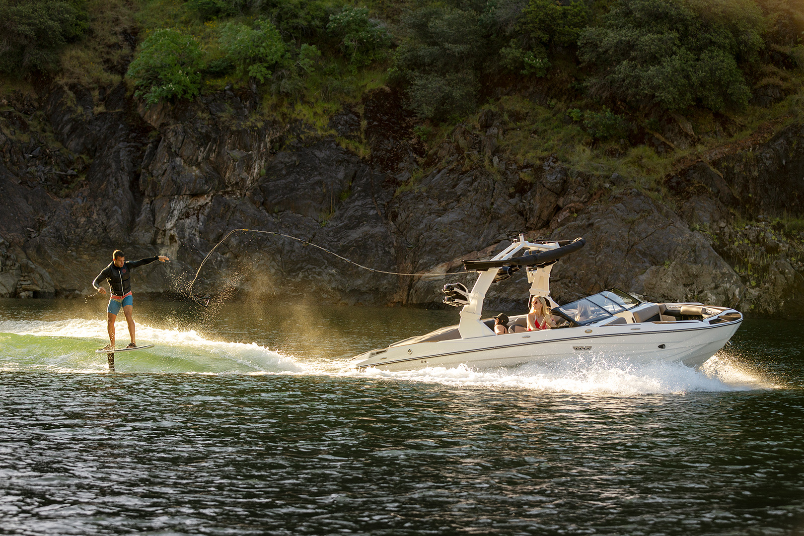 A person wakeboarding behind a white Supreme S220 motorboat on a lake near a rocky, tree-covered shore.