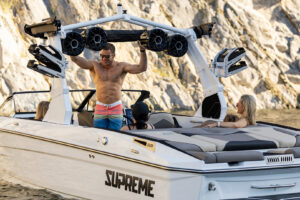 A man in swim trunks stands on a white boat labeled “SUPREME” with three other people seated onboard, cruising near rocky cliffs and sparkling water aboard the impressive Supreme Towboats S220.