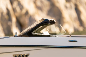A close-up side view of a sleek white Supreme S220 boat with a black windshield, showing chrome details and part of the control panel, set against a blurred rocky background.