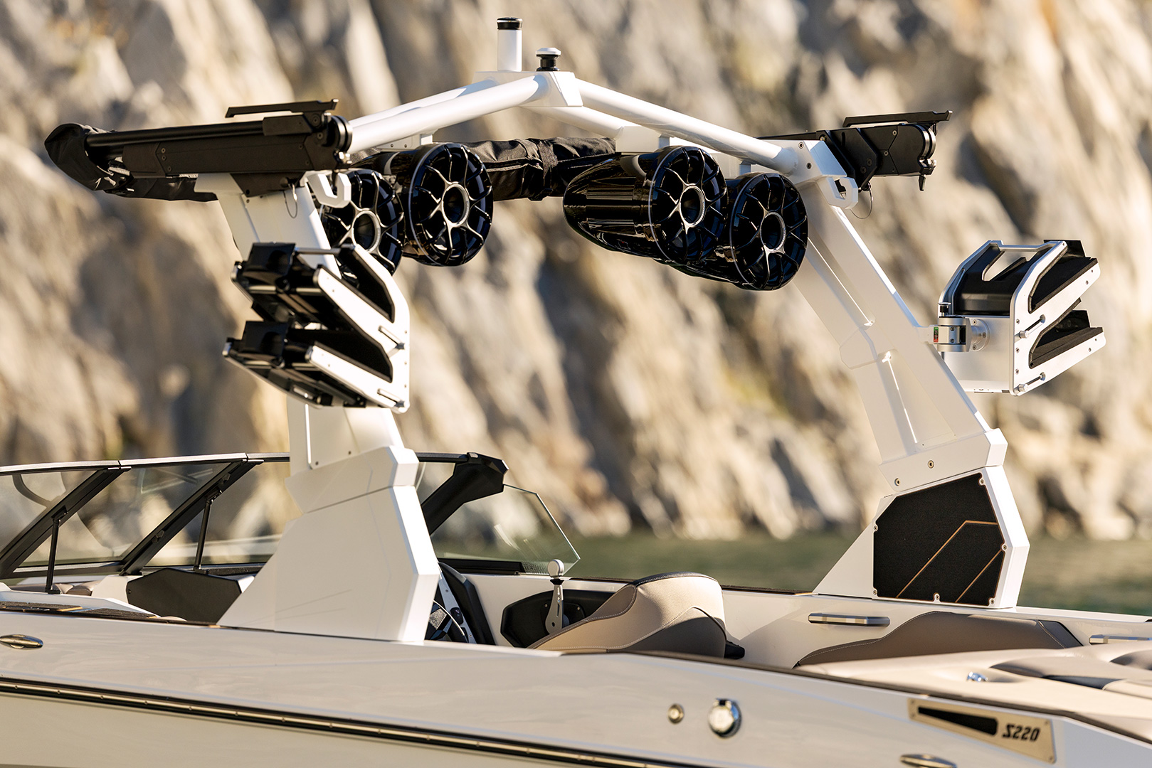 Close-up of a modern Supreme S220 motorboat with white and black wakeboard tower, multiple mounted speakers, and racks for wakeboards, set against a rocky shoreline background.