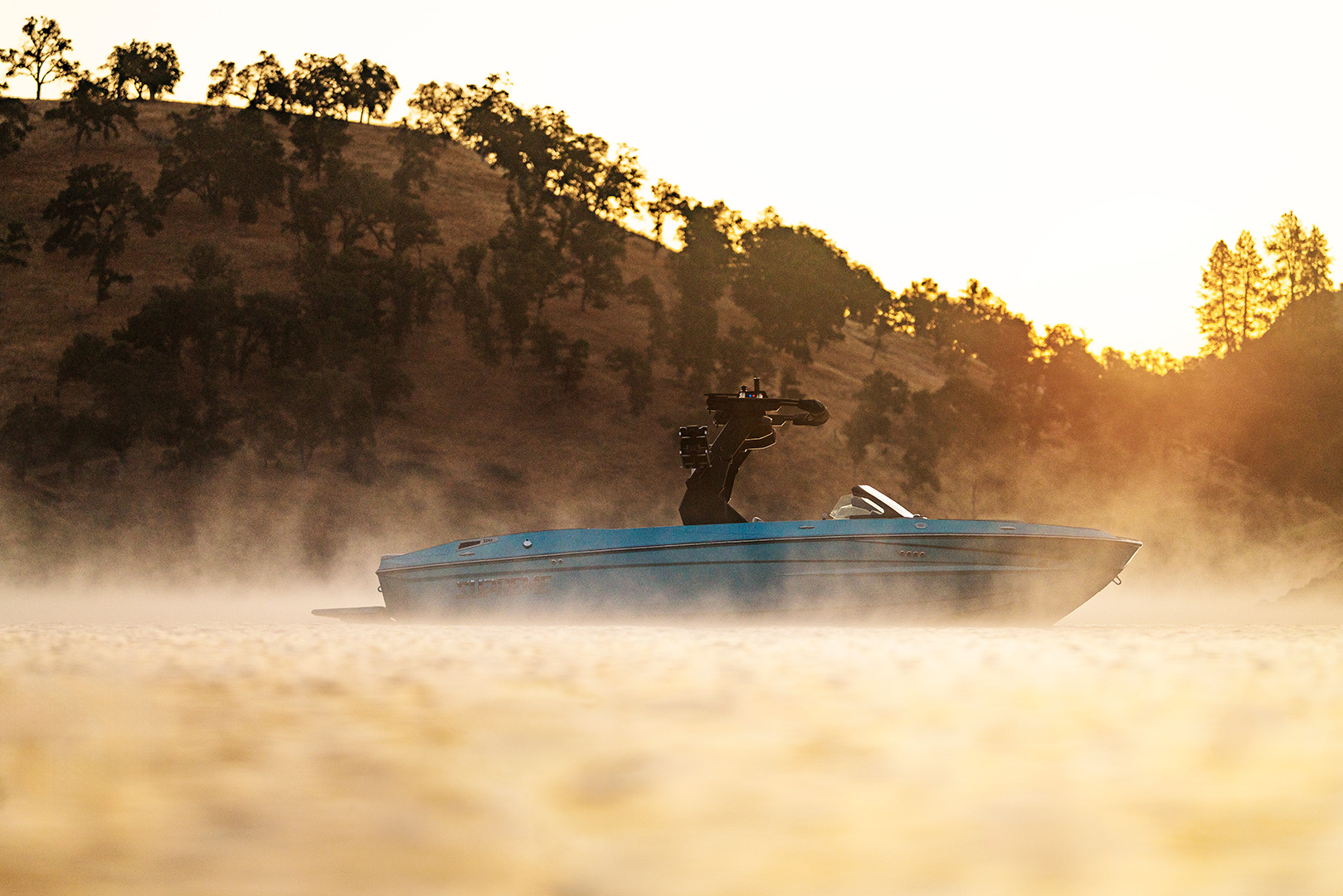 A blue Supreme S240 speedboat travels across a misty lake at sunrise, with a mounted camera on the boat. Tree-covered hills are visible in the background.