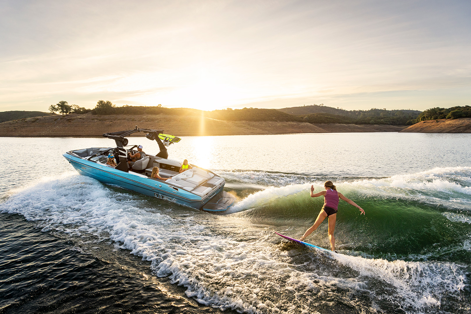 A person wake surfs behind a blue Supreme S240 motorboat on a calm lake at sunset, with gentle waves and distant hills in the background.
