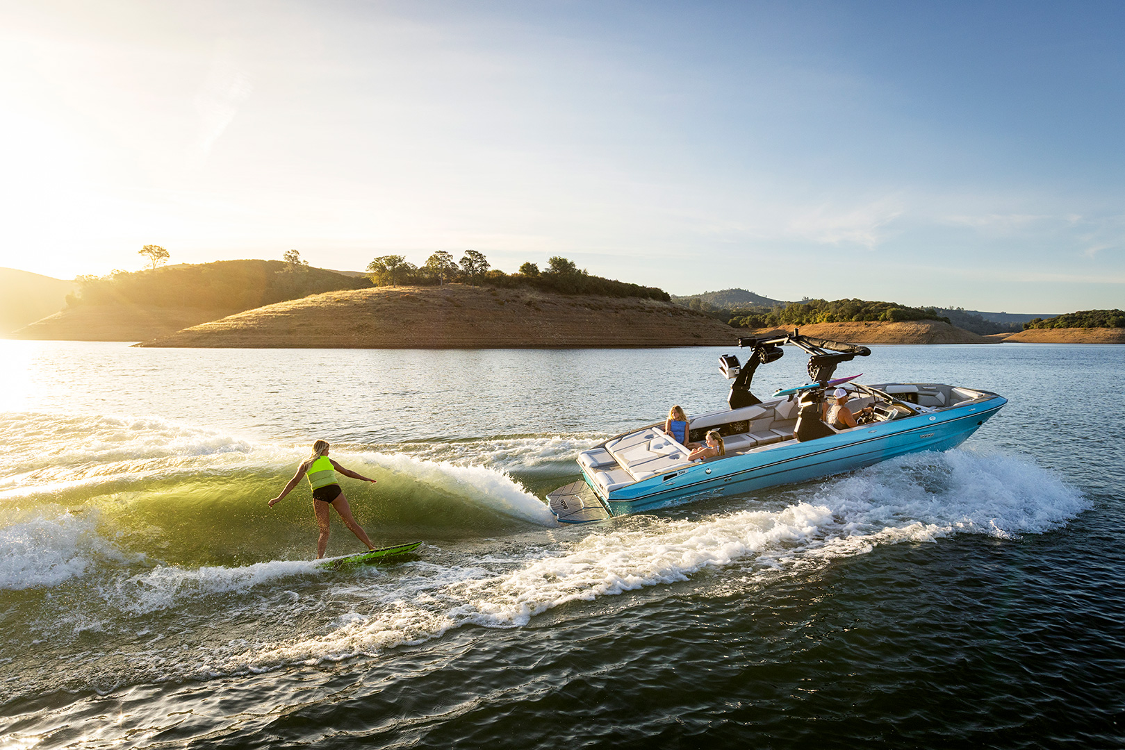 A person wake surfs behind a blue Supreme Boats S240 speedboat on a lake with low, grassy hills in the background during sunset.