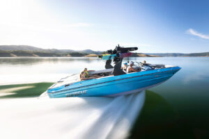 A blue Supreme Boats S240 speedboat with several people onboard moves quickly across a calm lake, creating a large wake, with hills and a clear sky in the background.