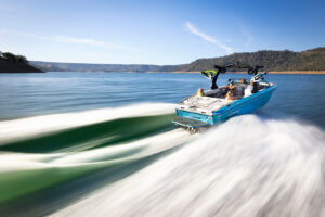 A blue Supreme S240 motorboat speeds across a lake, leaving white waves behind. Two people are sitting inside as the boat moves toward the open water and distant forested hills.