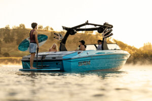 Four people are on a blue Supreme Boats S240 wakeboard boat in calm water at sunset; one person stands holding a wakeboard, while three others sit or lounge on the boat.