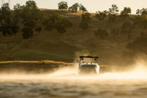 A Supreme S240 motorboat speeds across a lake, creating a spray of water, with a grassy hillside and scattered trees in the background.