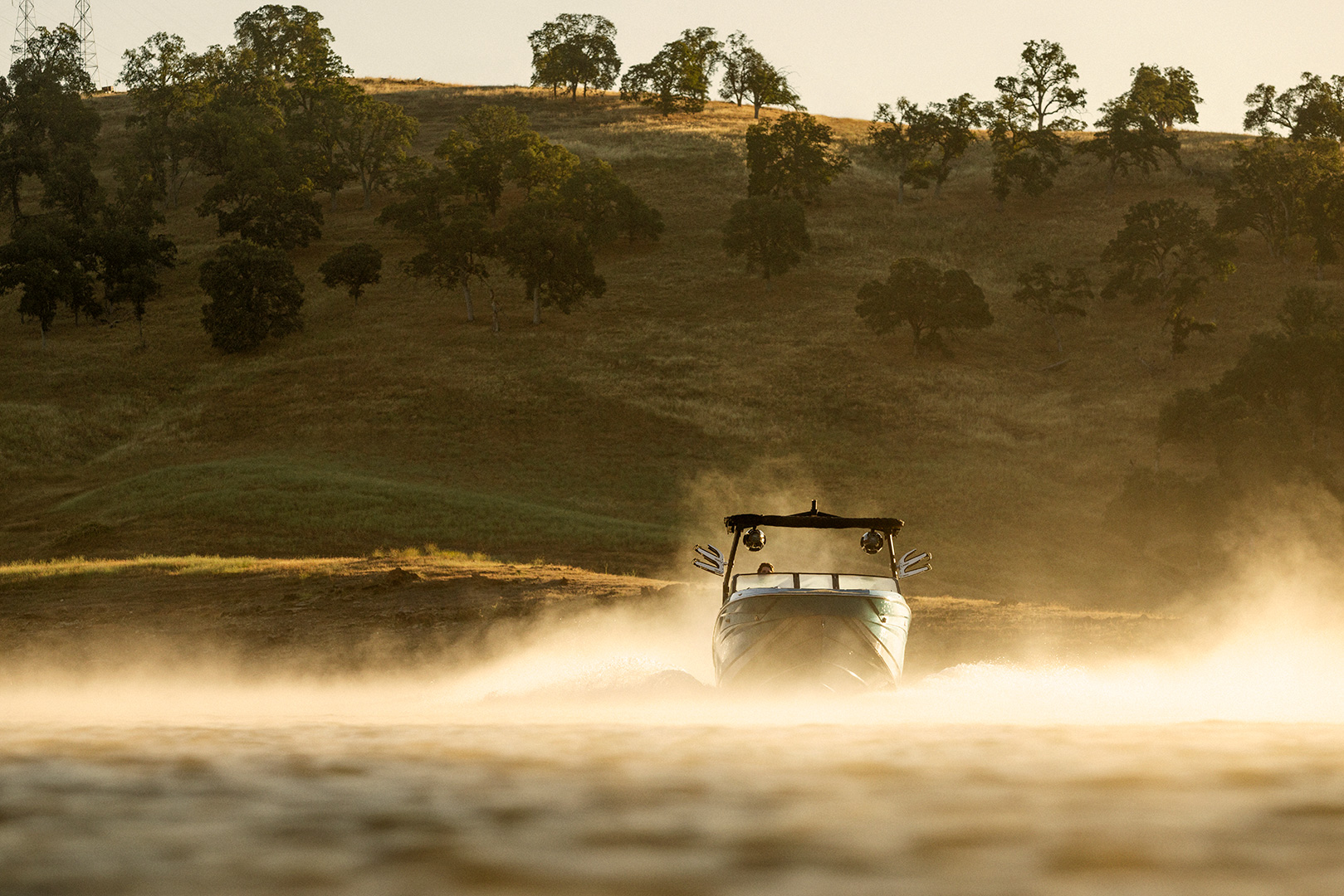 A Supreme S240 motorboat speeds across a lake, creating a spray of water, with a grassy hillside and scattered trees in the background.