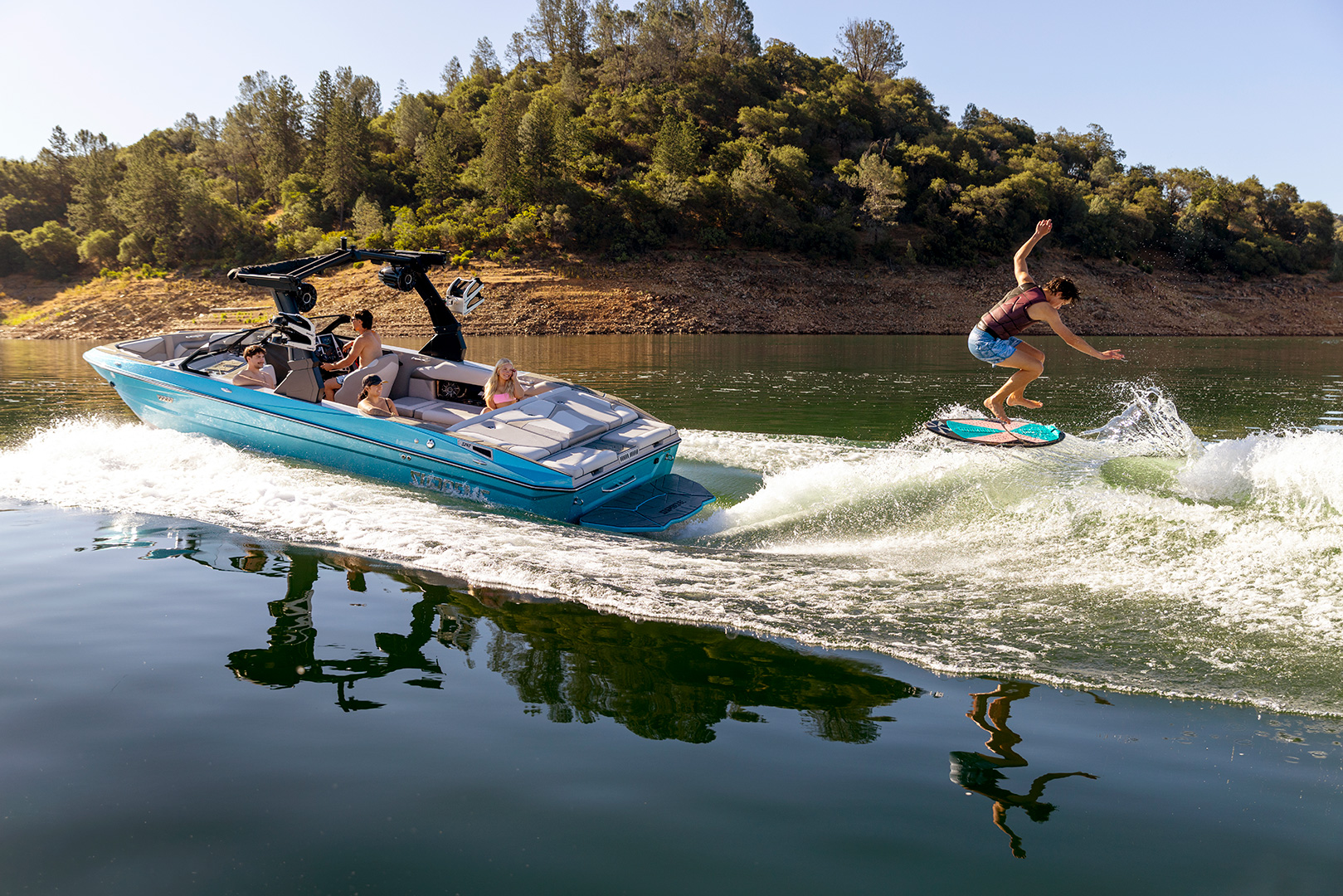 A person is wakesurfing behind a blue Supreme Boats S240 motorboat with three people relaxing on board, on a calm lake surrounded by wooded hills.