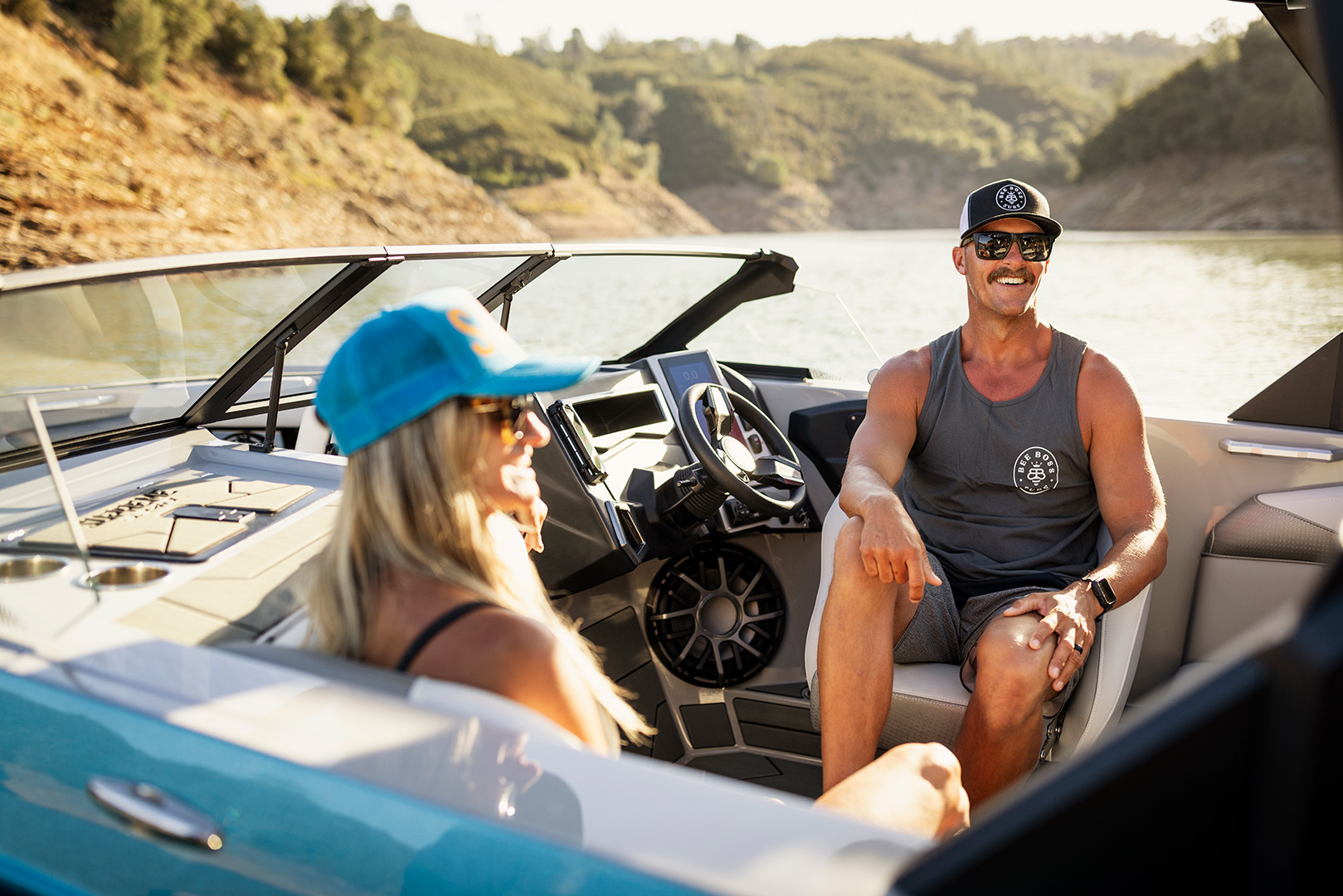 Two people wearing sunglasses and hats sit and relax in a Supreme Boats S240 speedboat on a sunny day, with water and greenery visible in the background.