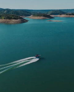 A Supreme Boats S240 speeds across a large blue lake, leaving a wake behind, with small tree-covered islands and distant hills in the background.